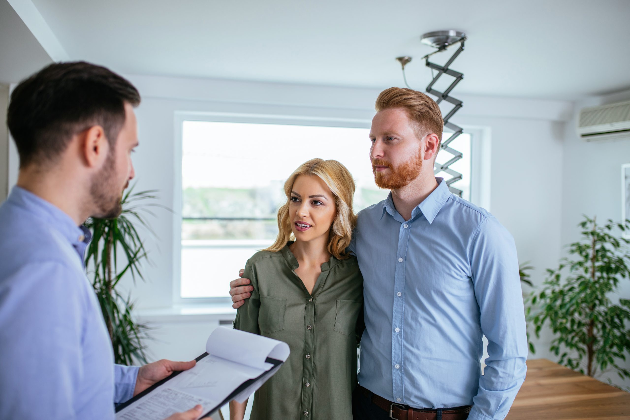 A couple talks with a real estate agent holding a clipboard inside a bright home.