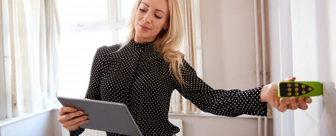 A woman in a bright room holds a tablet while using a handheld moisture meter against a wall, appearing to inspect or measure dampness.