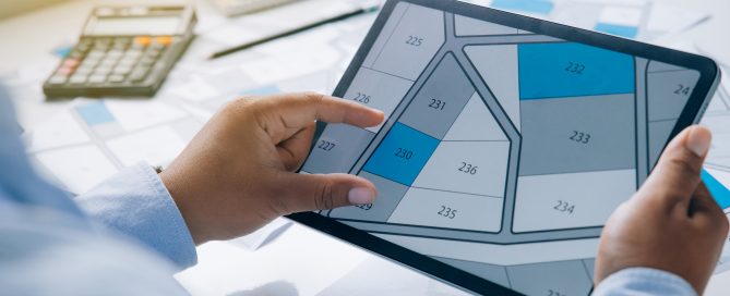 Person holding a tablet displaying a digital map of land plots with numbered sections, while a calculator and papers with printed maps are on the table.
