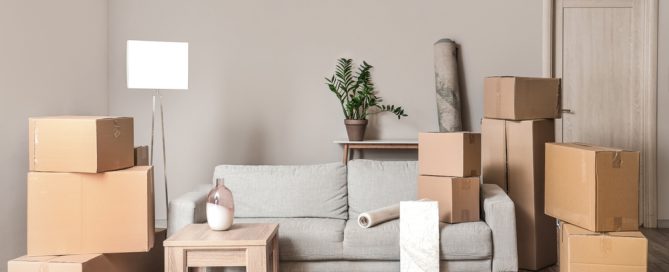 A bright living room with a sofa, small coffee table, floor lamp, and several stacked cardboard moving boxes, suggesting a home in the process of moving or unpacking.