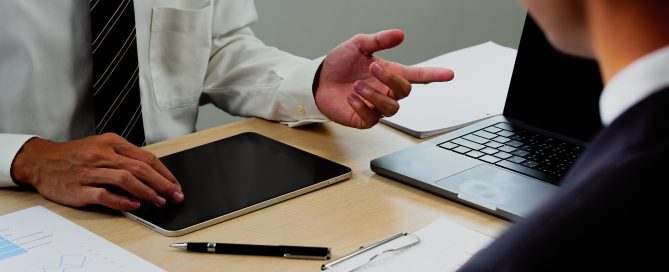 Two professionals seated at a desk during a business meeting, with one person gesturing while discussing data beside a tablet, laptop, and printed financial charts.