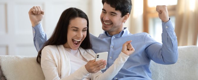Two people sitting on a sofa indoors, both cheering while looking at a smartphone one of them is holding. They appear excited and are raising their arms in celebration.