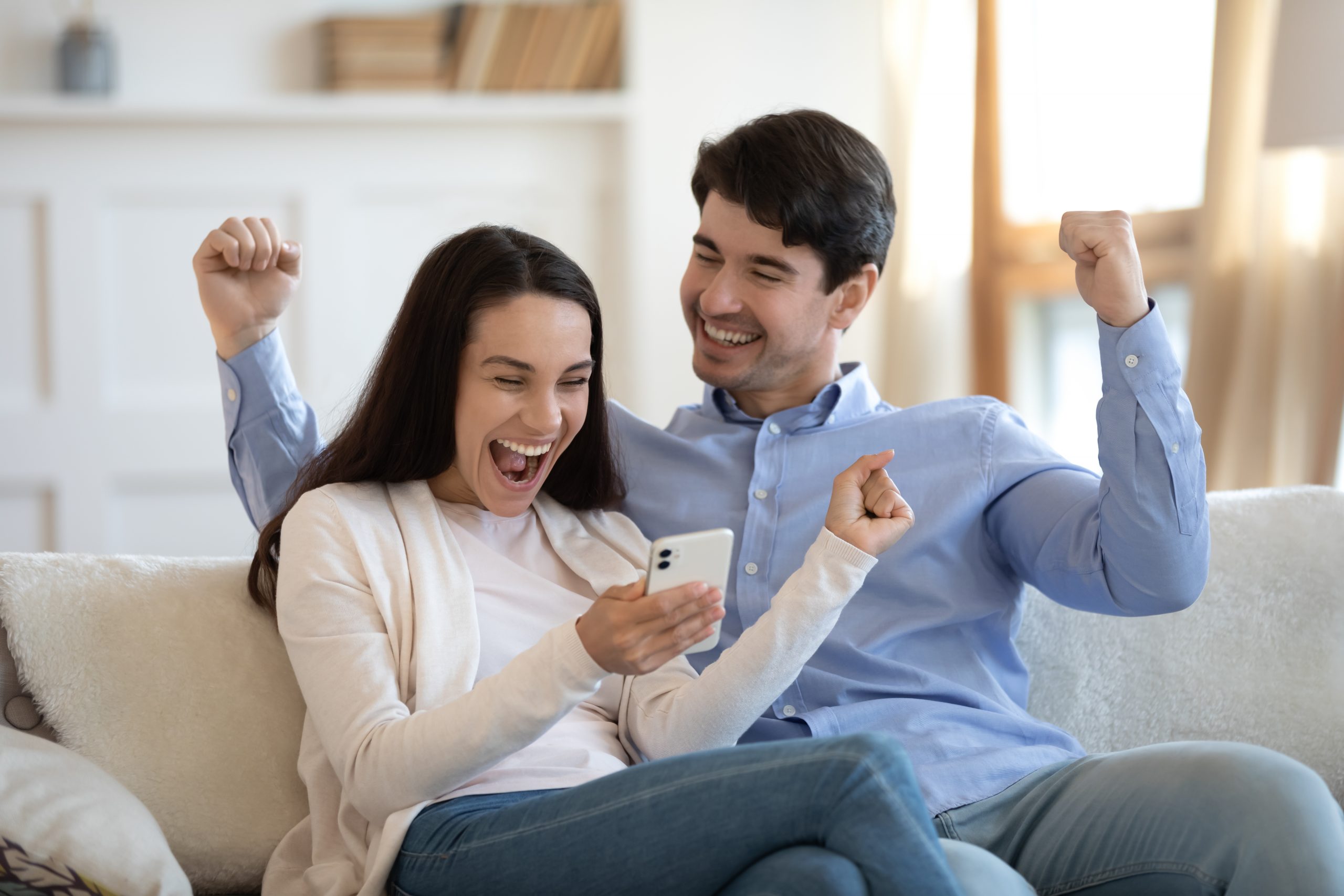 Two people sitting on a sofa indoors, both cheering while looking at a smartphone one of them is holding. They appear excited and are raising their arms in celebration.