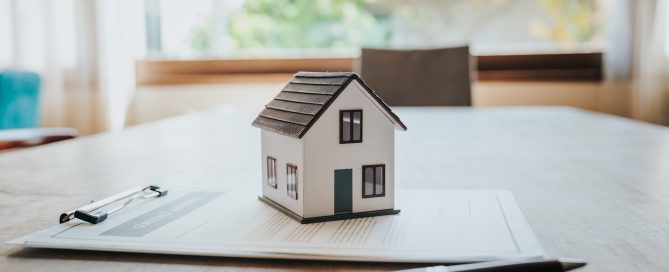 A small model house sits on top of property contract documents on a wooden table, with a pen and clipboard nearby. The image is softly lit by natural light from a window in the background, symbolising home ownership, real estate transactions, or mortgage agreements.