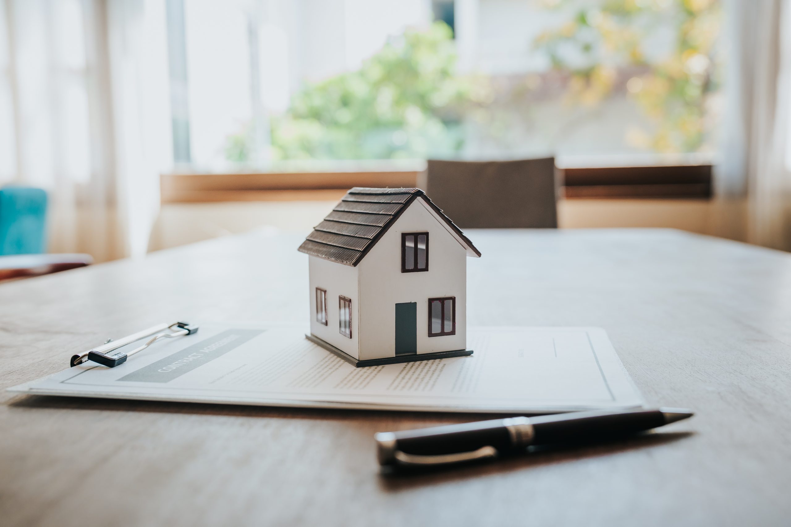 A small model house sits on top of property contract documents on a wooden table, with a pen and clipboard nearby. The image is softly lit by natural light from a window in the background, symbolising home ownership, real estate transactions, or mortgage agreements.