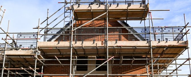 A brick building covered in multi‑level metal scaffolding during construction or renovation. Wooden planks form walkways across the structure, with safety railings surrounding each level. The building has a triangular gable roof partially visible behind the scaffolding, all set against a bright blue sky.