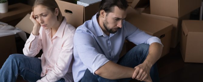 Two people sit on the floor with their backs against each other in a room filled with cardboard moving boxes. Both are dressed casually in button-up shirts and jeans. Several packed boxes, storage shelves, and household items are visible in the background, suggesting a home relocation or moving process.