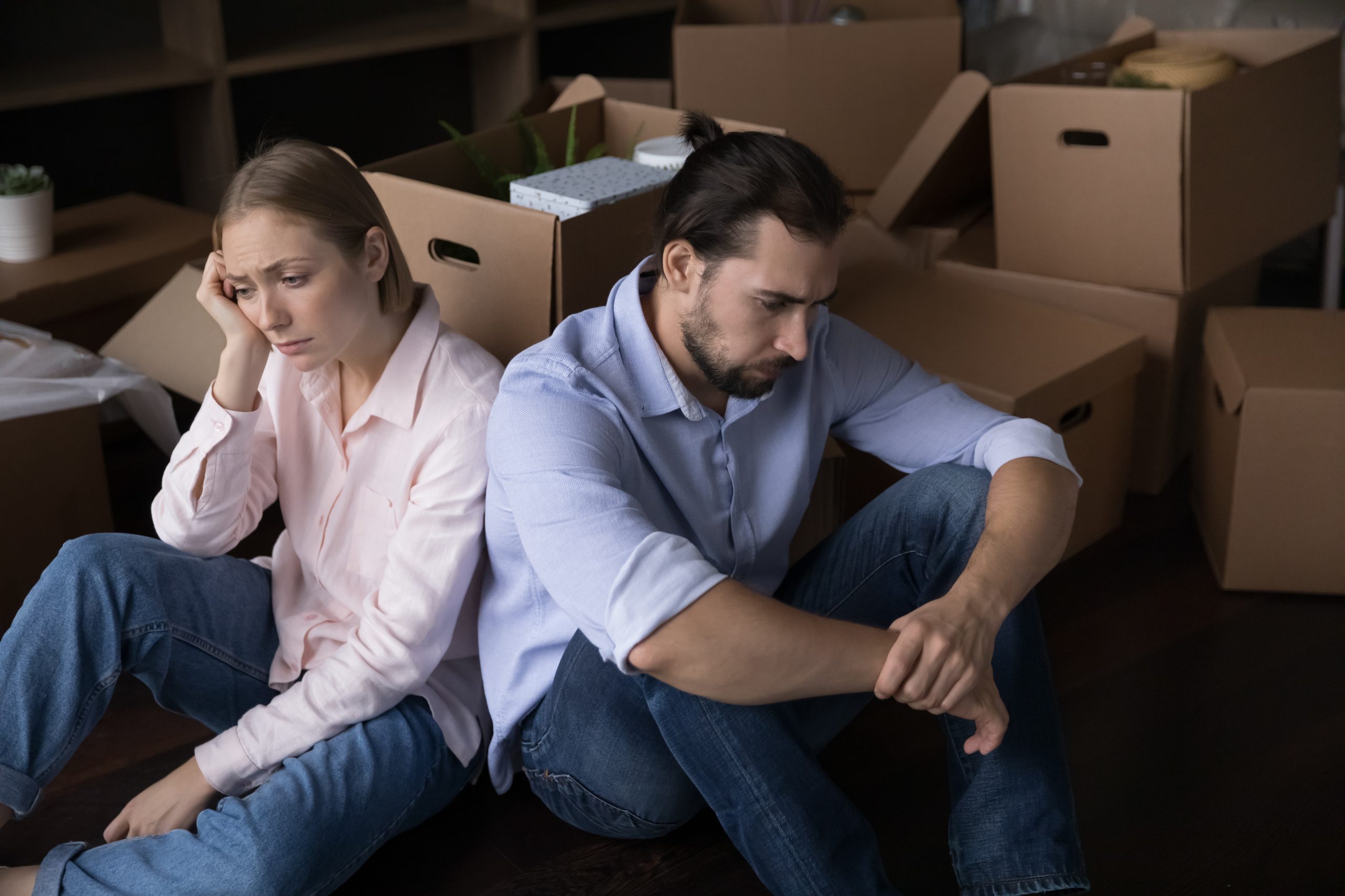 Two people sit on the floor with their backs against each other in a room filled with cardboard moving boxes. Both are dressed casually in button-up shirts and jeans. Several packed boxes, storage shelves, and household items are visible in the background, suggesting a home relocation or moving process.