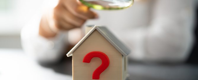 Close-up of a small model house with a large red question mark on the front, while a person in the background examines it with a magnifying glass. The image symbolizes property valuation, due diligence, and uncovering hidden costs or potential issues when purchasing a home.