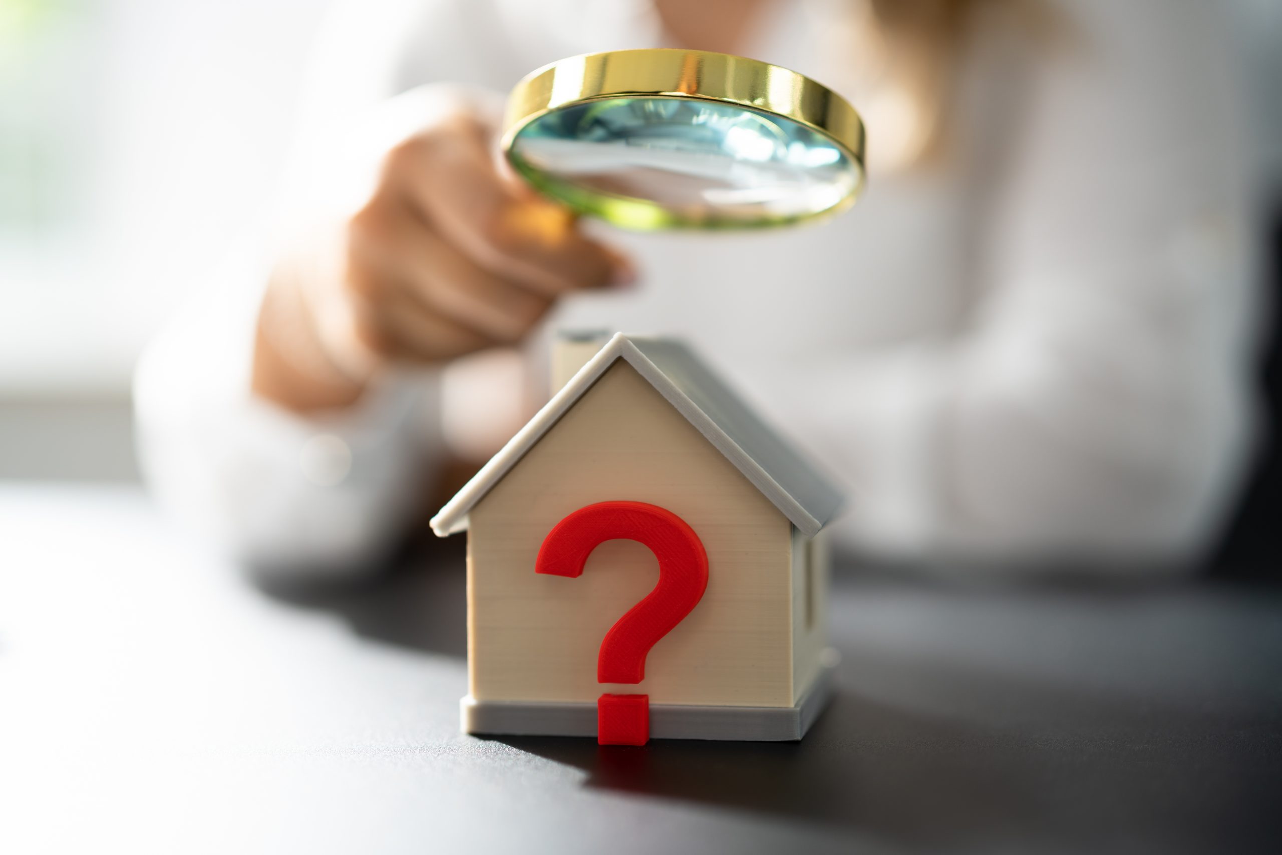 Close-up of a small model house with a large red question mark on the front, while a person in the background examines it with a magnifying glass. The image symbolizes property valuation, due diligence, and uncovering hidden costs or potential issues when purchasing a home.