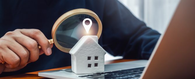 Close-up of a white model house placed on a laptop keyboard while a person holds a magnifying glass over it. A location pin icon appears above the house, symbolising online property search, real estate research, location-based services, or property valuation.