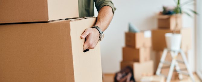 Close-up of a person carrying two large cardboard moving boxes in a home. Additional packed boxes, a plant on a stand, and household items are visible in the background, suggesting a house move or relocation.