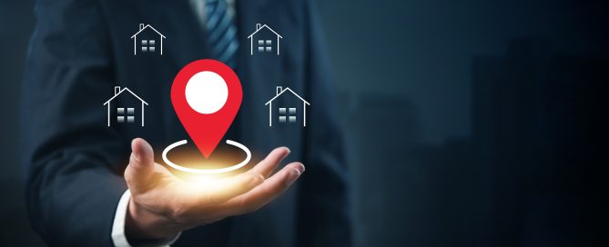 A business professional extends an open hand displaying a glowing red location pin icon, surrounded by four house icons. The digital graphic represents property location, real estate mapping, geolocation services, or local property search technology against a dark blue background.