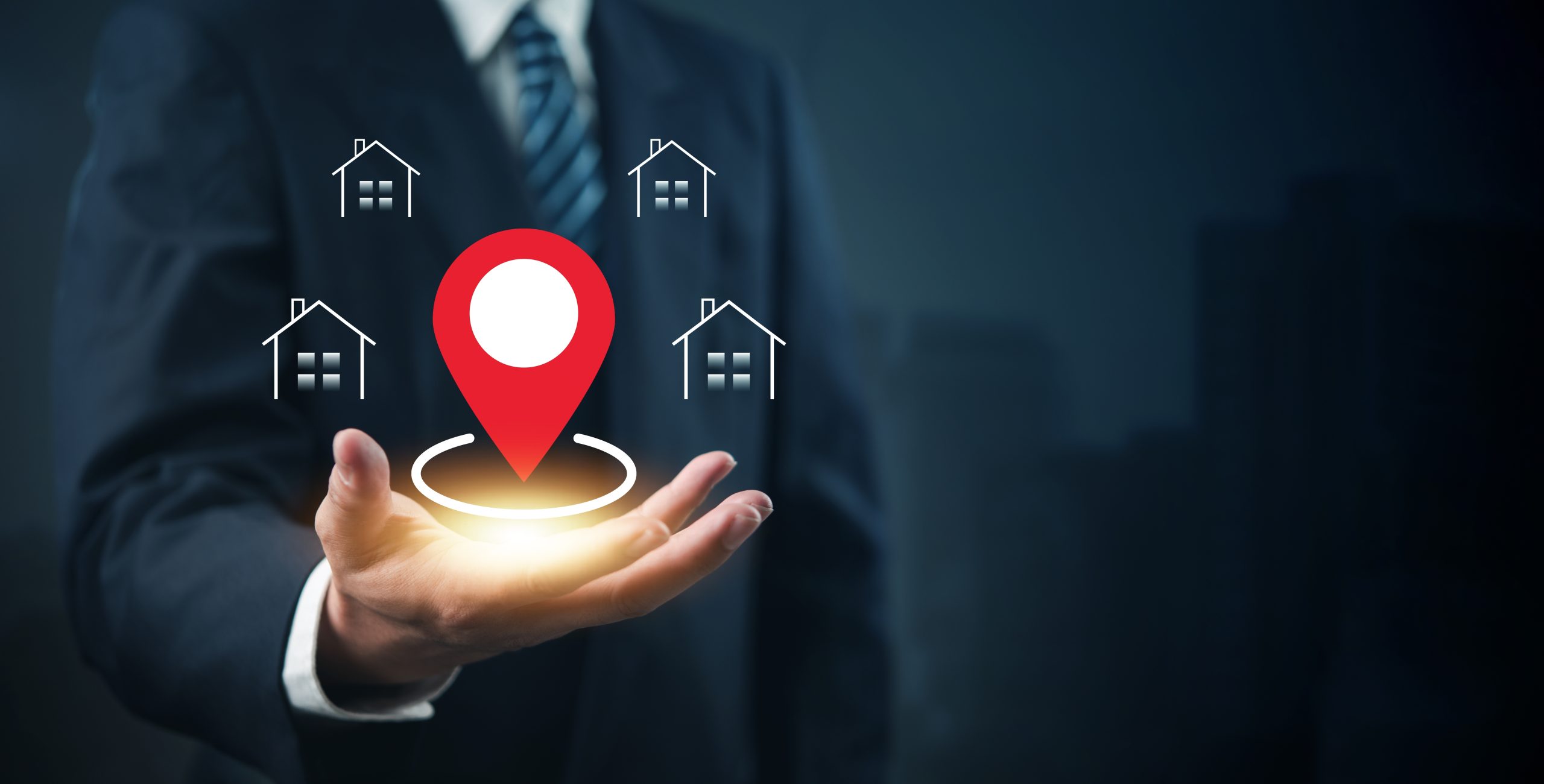 A business professional extends an open hand displaying a glowing red location pin icon, surrounded by four house icons. The digital graphic represents property location, real estate mapping, geolocation services, or local property search technology against a dark blue background.