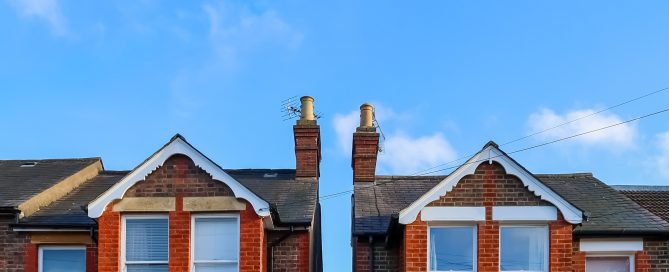 Front view of two adjoining Victorian-style red-brick terraced houses with white-trimmed bay windows and decorative gables. Twin chimneys rise above slate roofs, with a narrow passageway between the properties. The houses are illuminated by bright daylight beneath a clear blue sky with a few scattered clouds.