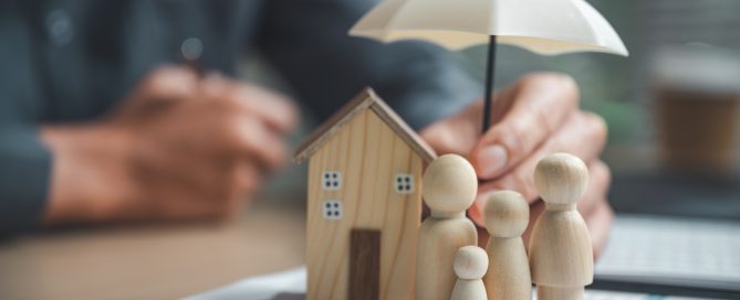 Wooden model figures representing a family stand beside a miniature wooden house under a white umbrella. The models are placed on financial documents, while a person in the background holds the umbrella above them, symbolizing home insurance, financial security, and family protection.