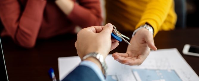 Close-up of a person handing a set of house keys to another person across a desk, with property floor plans and documents visible in the foreground and two people seated in the background. The image represents a home purchase, property transaction, or new home ownership.