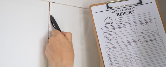 Close-up of a property inspector examining a tiled interior wall with a handheld tool while holding a home inspection report on a clipboard. The inspection focuses on assessing wall condition, cracks, or defects as part of a residential property survey.