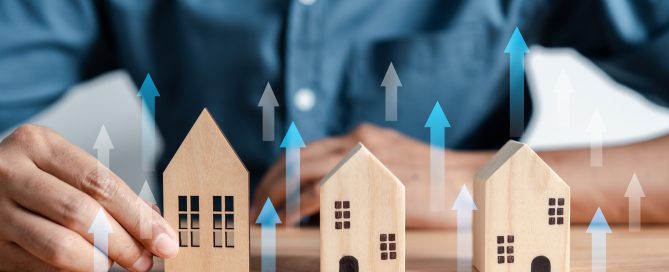 Three wooden model houses arranged on a table, with a person's hand beside the largest house. Multiple translucent blue and white arrows point upward around the houses, symbolising rising property values, house prices, real estate growth, or increased market demand. The background shows a person in a blue shirt out of focus.