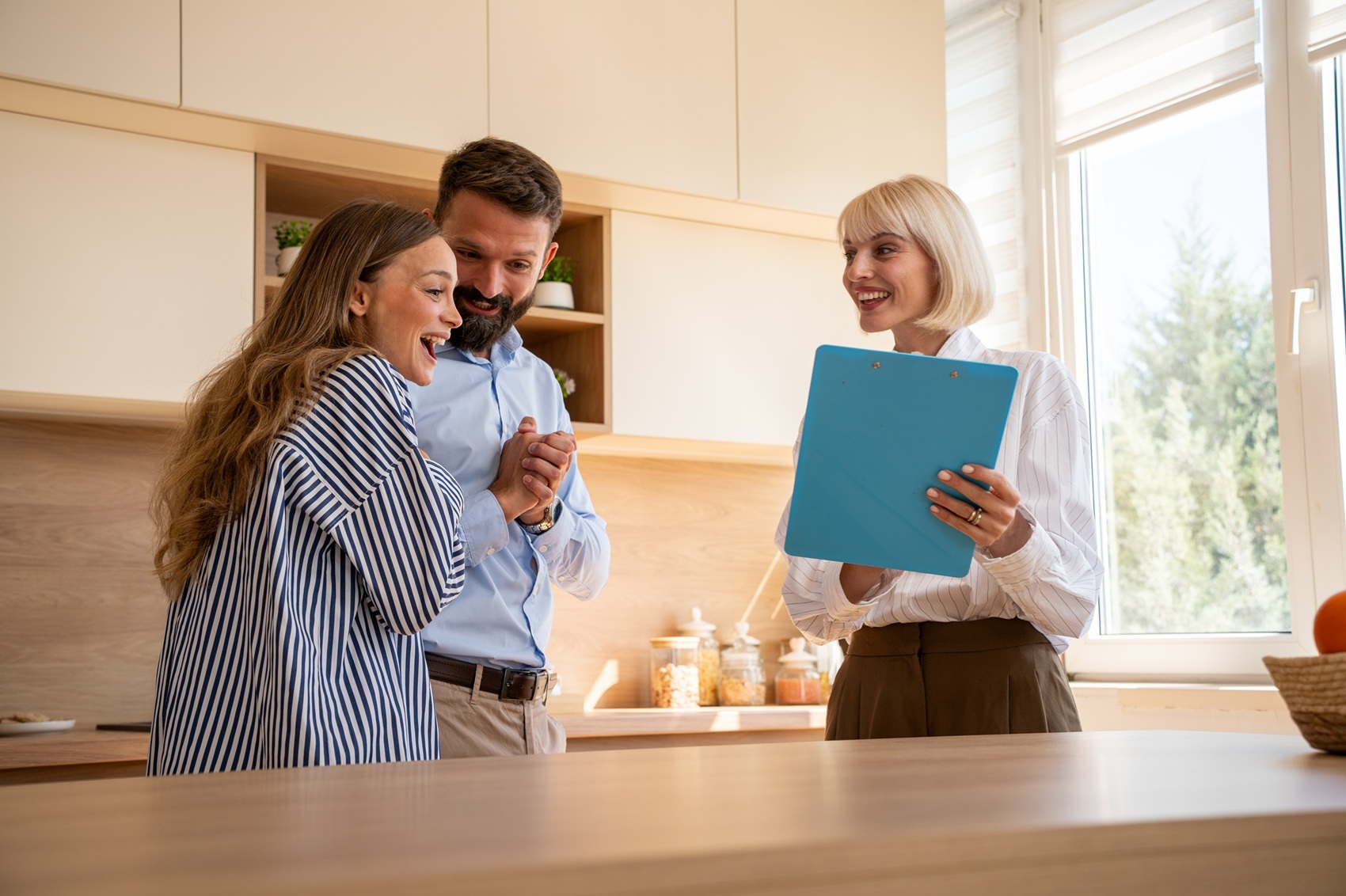 Estate agent holding a clipboard while meeting with two prospective homebuyers in a modern kitchen. The individuals stand beside a kitchen island as the estate agent discusses property details during a home viewing. Bright natural light streams through a large window, highlighting the contemporary cabinetry and clean interior design.