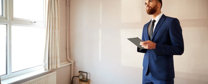 Person in a blue business suit holding a digital tablet while inspecting an unfurnished room. The room features a large window with curtains, a radiator, exposed pipework, and a gas meter, suggesting a residential property valuation or survey is being carried out.