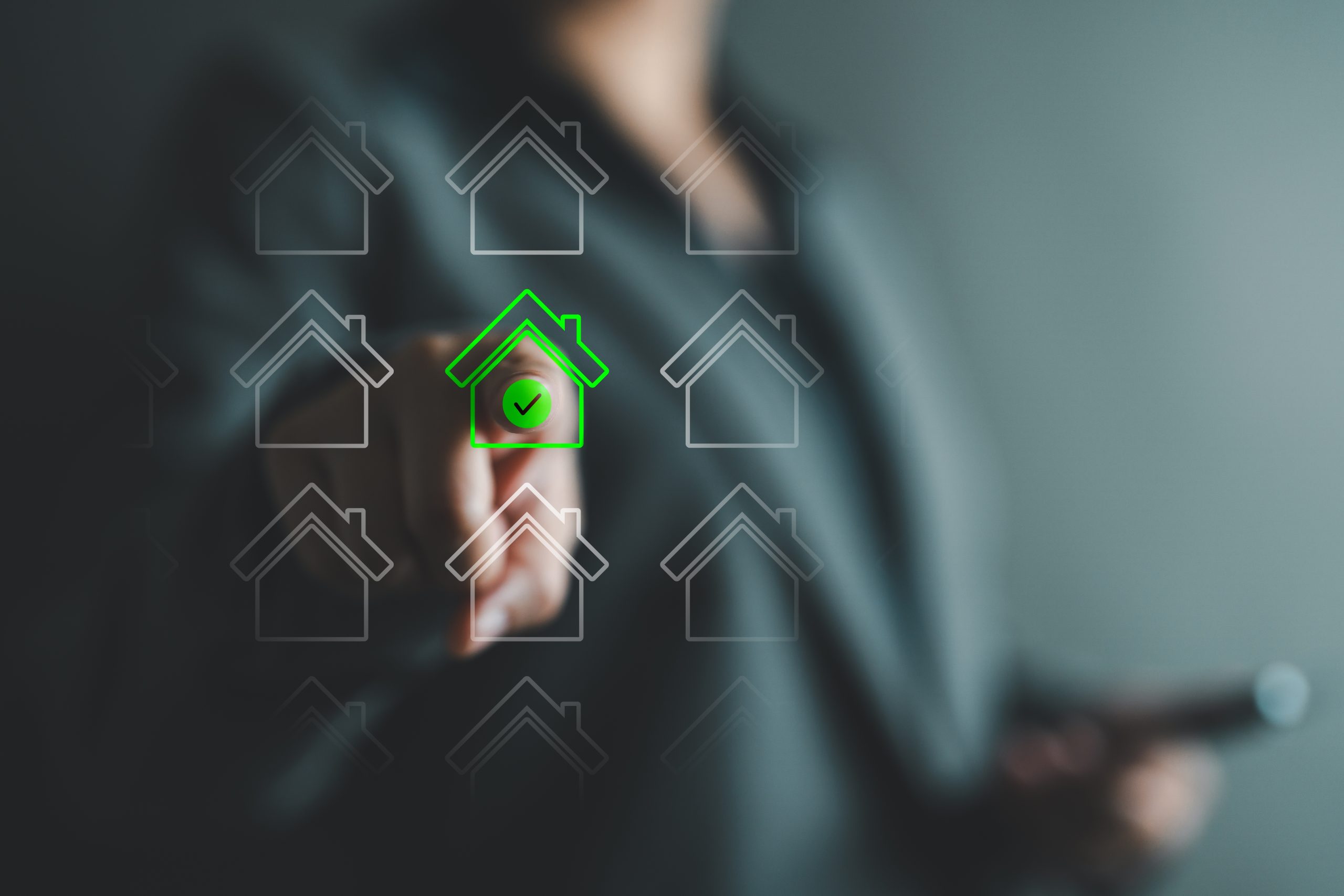 A close-up image of a hand interacting with a digital interface displaying multiple house icons. One house icon in the center is highlighted in bright green with a green checkmark, indicating an approved or selected property. The surrounding house icons are semi-transparent and greyed out against a blurred background.